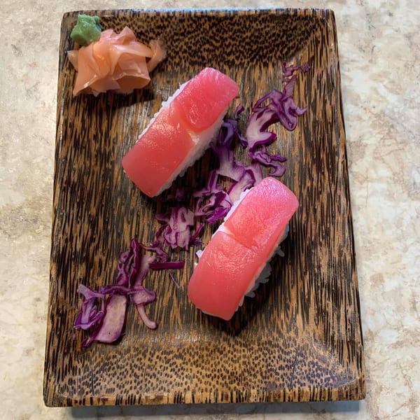 Two pieces of tuna nigiri sushi with pickled ginger, wasabi, and red cabbage on a wooden plate.