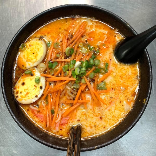 Spicy ramen with sliced carrots, green onions, and two halves of a boiled egg in a black bowl.